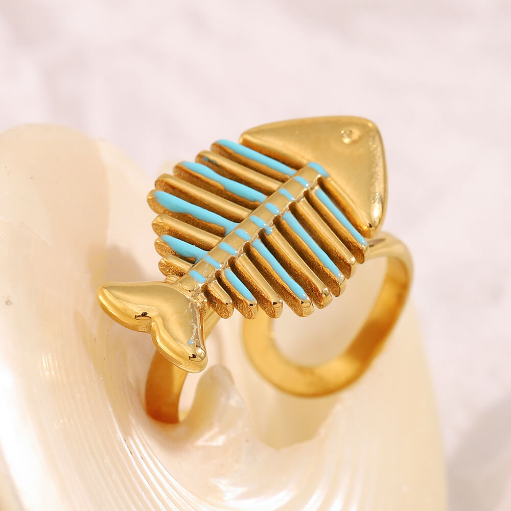 Gold fish-shaped ring with turquoise accents on a beige background.