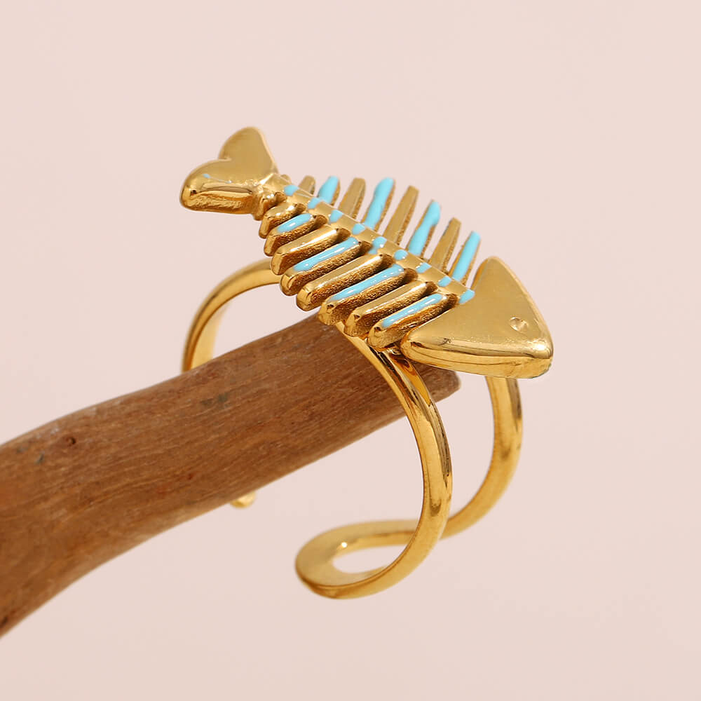 Gold fish bone-shaped ring with blue accents on a beige background.