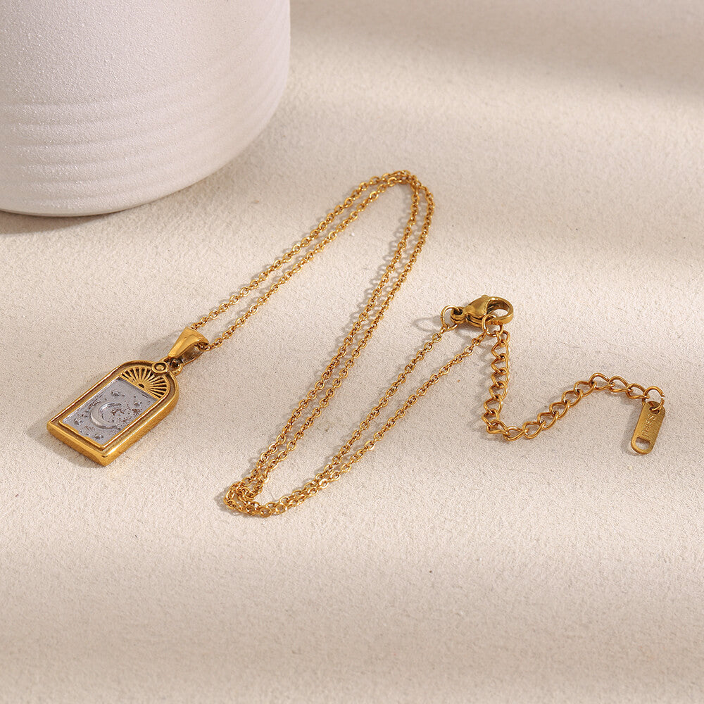 Gold celestial necklace with a rectangular pendant on a light beige surface.